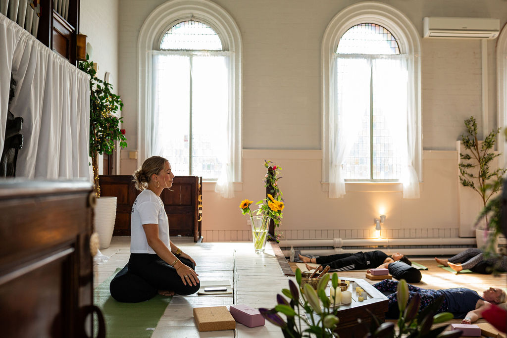 A teacher sitting quietly at the front of the studio beneath tall arched windows
